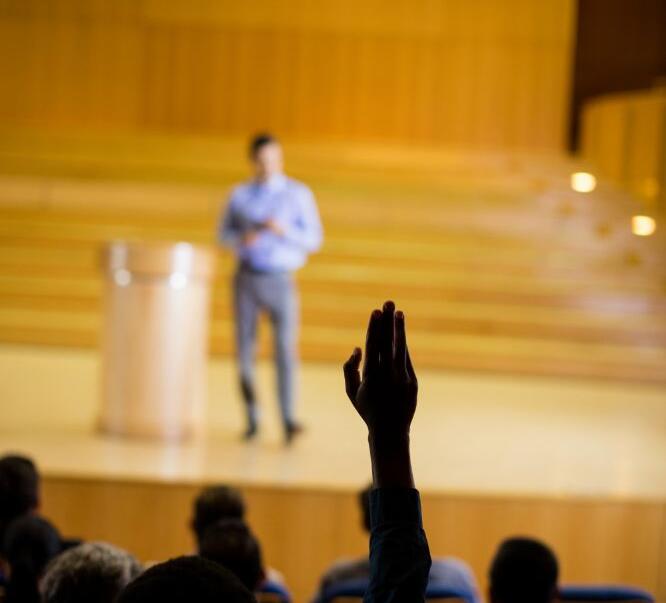 Expositor en auditorio. Persona entre el público levanta la mano para hacer una pregunta